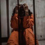 Person in orange jumpsuit sitting behind bars, handcuffed.