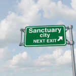 Illinois Town Grieves — Sanctuary Laws STRIKE AGAIN! Green road sign saying sanctuary city next exit