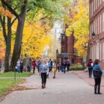 Students walking on a campus path surrounded by autumn trees and brick buildings