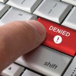 A finger pressing a red DENIED button on a computer keyboard