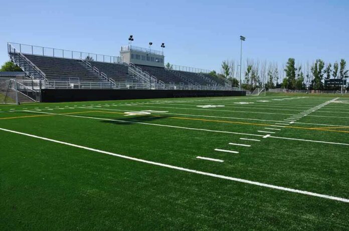 A football field with artificial turf and empty bleachers under a clear blue sky