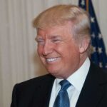 Trump Wax Figure ATTACKED – Shocking RESPONSE! A smiling man in formal attire with an American flag in the background