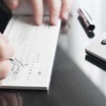 SCANDAL — The Dead Still Cashing Housing Checks Person writing a check on a reflective surface with a calculator nearby