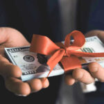 Biden’s Parting Gift—MONSTER WHITE ELEPHANT Hands holding stack of hundred-dollar bills with ribbon.