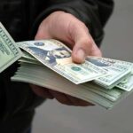 Millions Still Paid Out Amid Shocking Fraud Scheme A hand holding a stack of hundred dollar bills with one bill prominently displayed