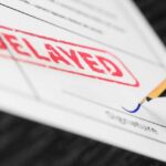 A close-up of a document with a red 'DELAYED' stamp and a fountain pen poised to sign