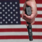 Riot Testifier’s Wild Power Play A hand holding a gun in front of an American flag