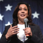Woman speaking with microphone, American flag background.