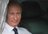 A smiling man in formal attire sitting inside a car with curtains