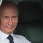A smiling man in formal attire sitting inside a car with curtains