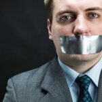 Man in business attire with duct tape over his mouth, conveying a message about silence