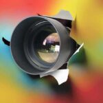 Camera lens emerging through torn colorful paper