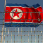 North Korea Signals Military Expansion in Factory Visit Barbed wire fence with North Korean flag behind.