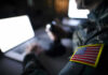 War Machine Runs On Musk’s Grok Soldier using laptop with US flag patch visible.
