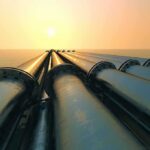 Shock Warning: Oil Chief Warns Crisis Has No Quick Fix Industrial pipes extending towards the horizon over water during sunset