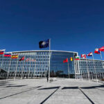 Trump Says ‘Finish the Job.’ Experts Say the Story Isn’t Over. Flags outside NATO headquarters building under clear blue sky.