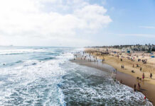 Vacations End: Deadly Illness Claims Cast Shadow Crowded beach with waves and people enjoying sun.