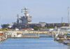 Bridge Bombings Mark Major Escalation in U.S.–Iran Conflict A large naval aircraft carrier docked in a harbor with smaller boats in the foreground