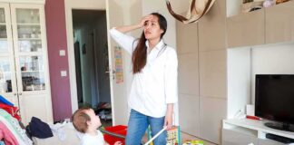 Mother standing in messy living room with toddler on couch