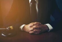 person in suit with hands clasped at a desk beside a gavel