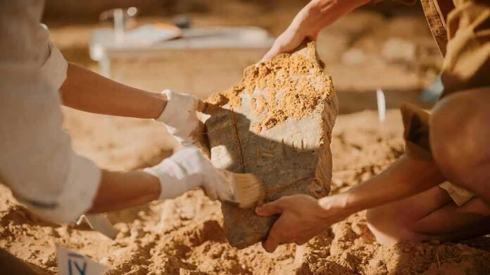 shutterstock_1955213146 Archaeologists cleaning a large stone artifact at a dig site