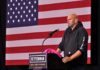 Man in hoodie speaking at a podium before a large U.S. flag