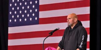 Man in hoodie speaking at a podium before a large U.S. flag