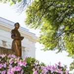 Bronze statue in a garden with pink flowers and a neoclassical building