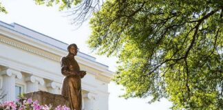 Bronze statue in a garden with pink flowers and a neoclassical building