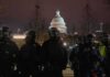 Riot police in protective gear stand before a domed capitol building at night
