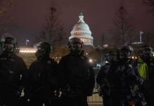 Riot police in protective gear stand before a domed capitol building at night