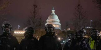Riot police in protective gear stand before a domed capitol building at night