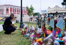 “Traitor” Chants Follow Mamdani at Vigil Police officer kneels at a memorial with flowers and crosses
