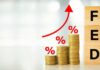 Massive Shake-Up: Viral Gold Forecast Sparks Questions Stacks of coins with rising red arrow and wooden blocks spelling FED